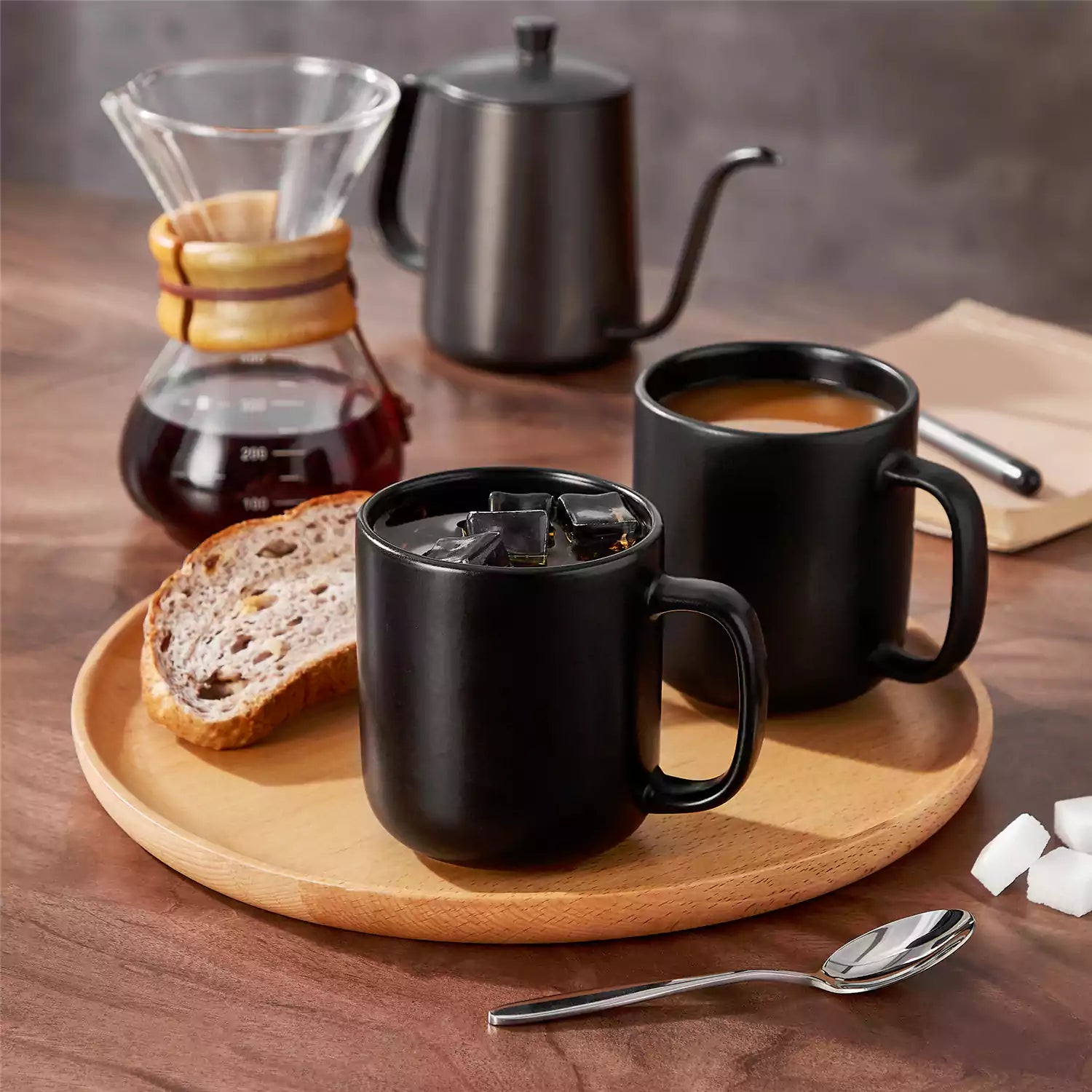 Playa 16oz stoneware mugs in black reactive glaze on a rustic wooden café table with coffee maker and pastries