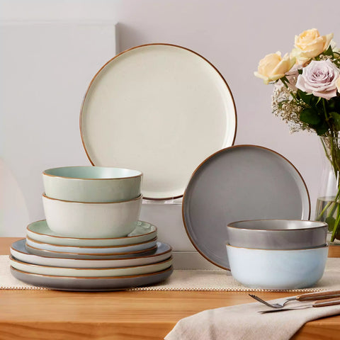 Assorted rustic stoneware dinnerware set with speckled - lipped-edge plates and bowls on a dining table with flowers and cutlery in a cozy modern kitchen-vancasso