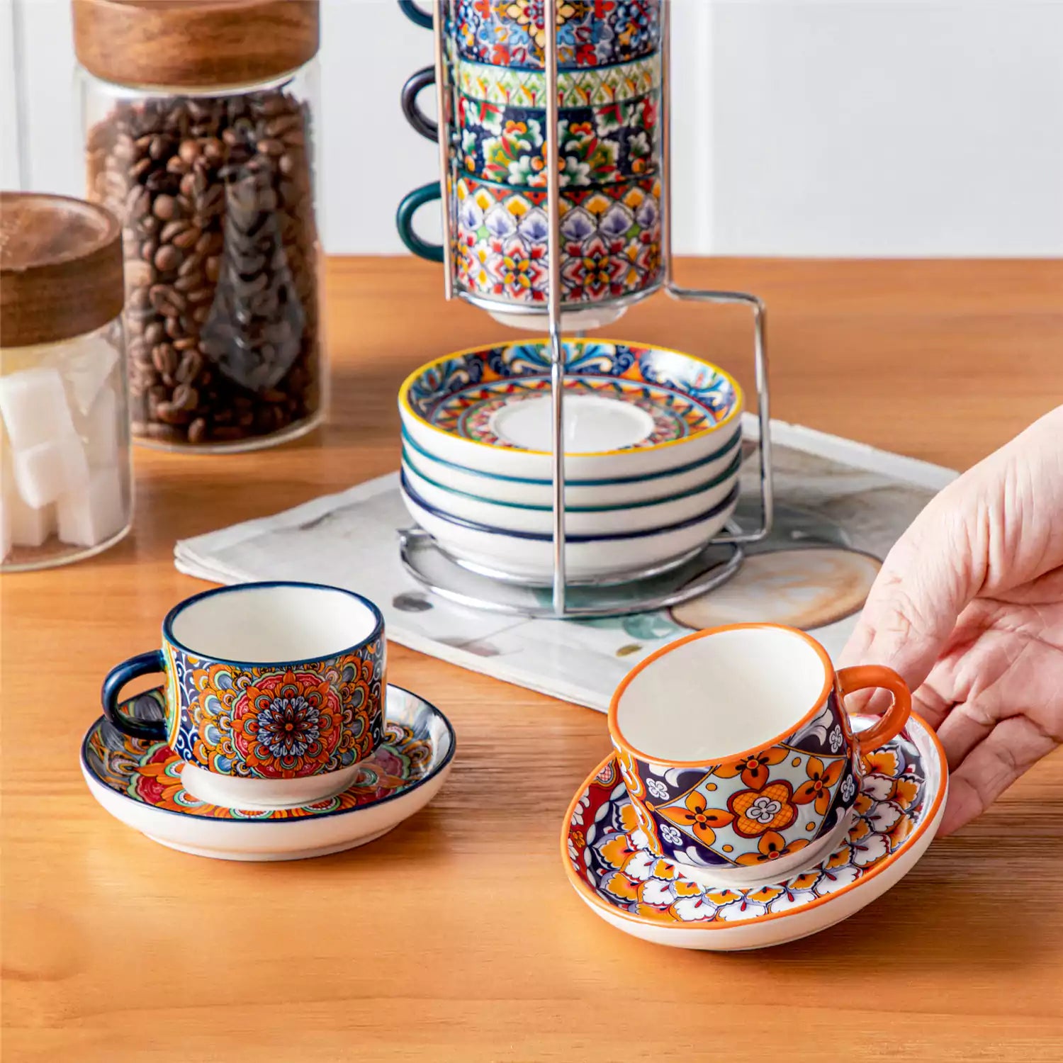 Simi 12 piece porcelain espresso set with 3oz cups and saucers in vibrant boho chic floral patterns on a black metal storage rack with a magazine and napkin on a wooden table